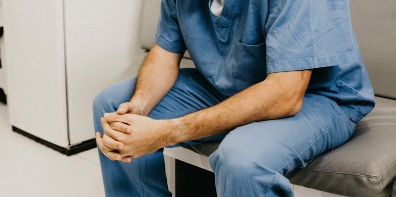 A male doctor in hospital attire sitting pensively, representing healthcare challenges.