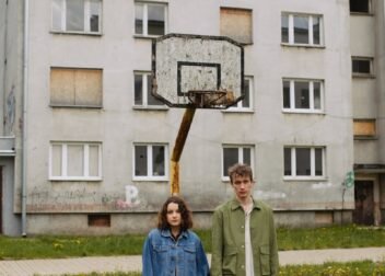 A young couple holding hands stands in front of an abandoned building with a rusty basketball hoop.