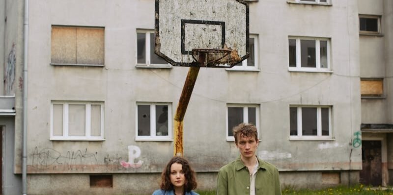 A young couple holding hands stands in front of an abandoned building with a rusty basketball hoop.