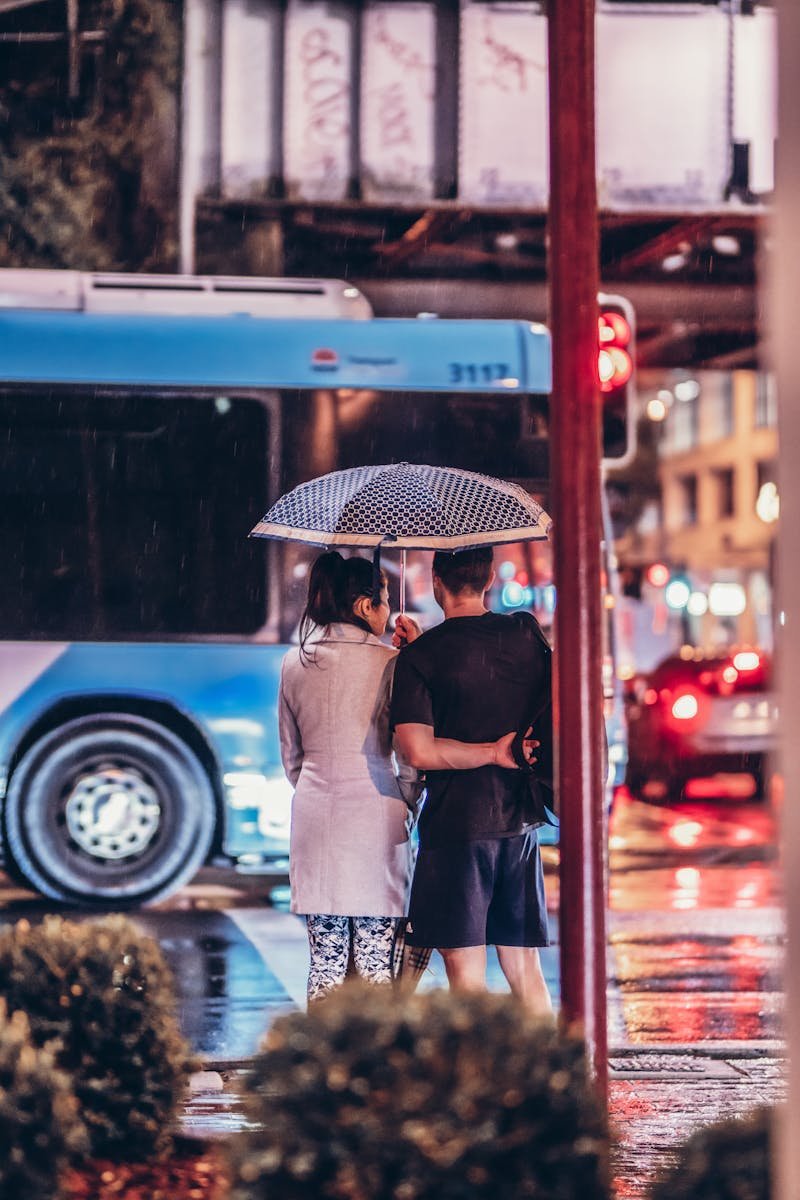 A couple stands under an umbrella, embracing on a rainy urban street at night.