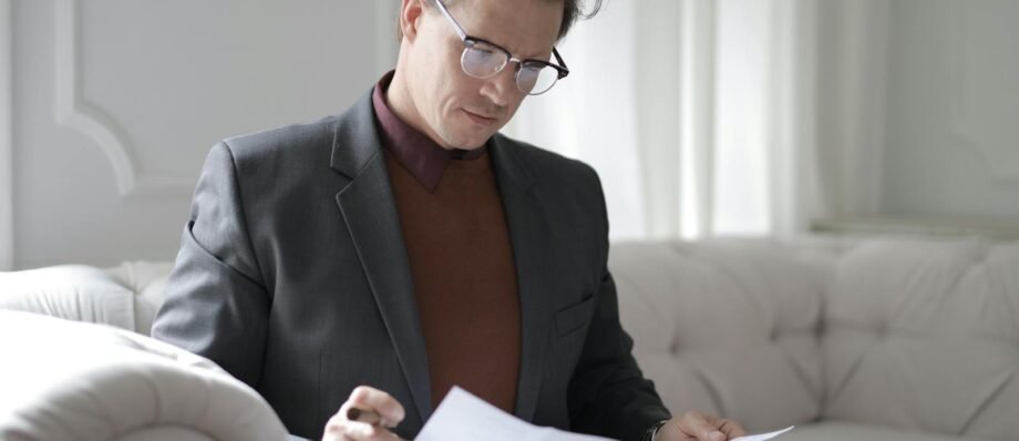 Elegant adult man in jacket and glasses looking through documents while sitting on white sofa in luxury room