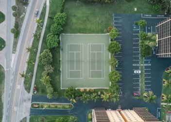 Overhead drone shot of a tennis court surrounded by greenery and parking in a cityscape.