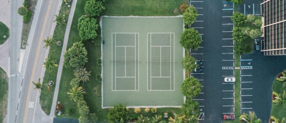 Overhead drone shot of a tennis court surrounded by greenery and parking in a cityscape.