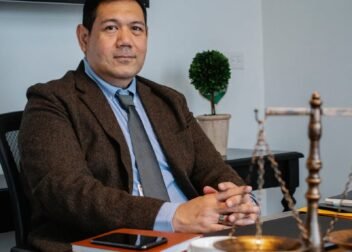 Middle aged ethnic legislator looking at camera and sitting at table with scales of justice in office