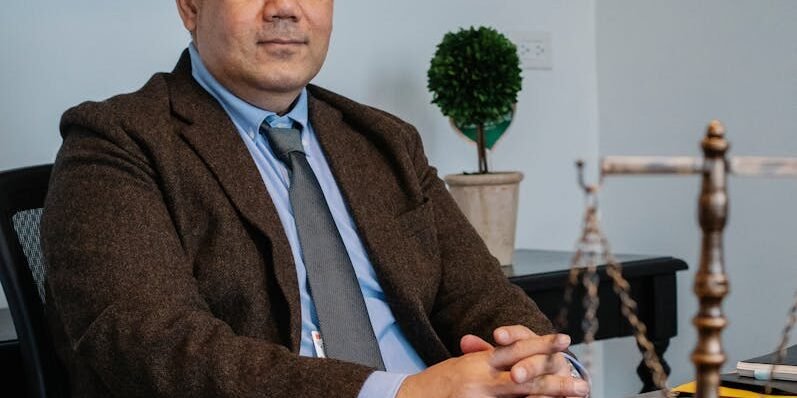 Middle aged ethnic legislator looking at camera and sitting at table with scales of justice in office