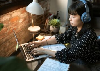 Focused woman wearing headphones, working on a laptop in a cozy home office setup with natural light.