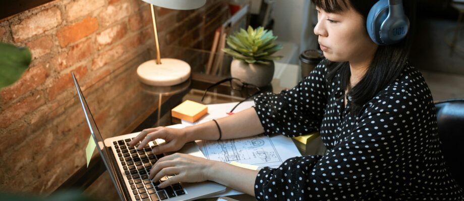 Focused woman wearing headphones, working on a laptop in a cozy home office setup with natural light.