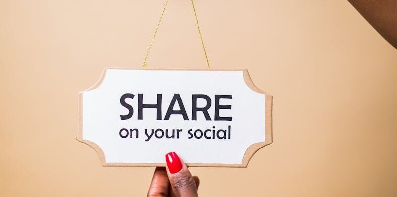 Close-up of a person's hand holding a 'Share on your social' sign against a beige background.