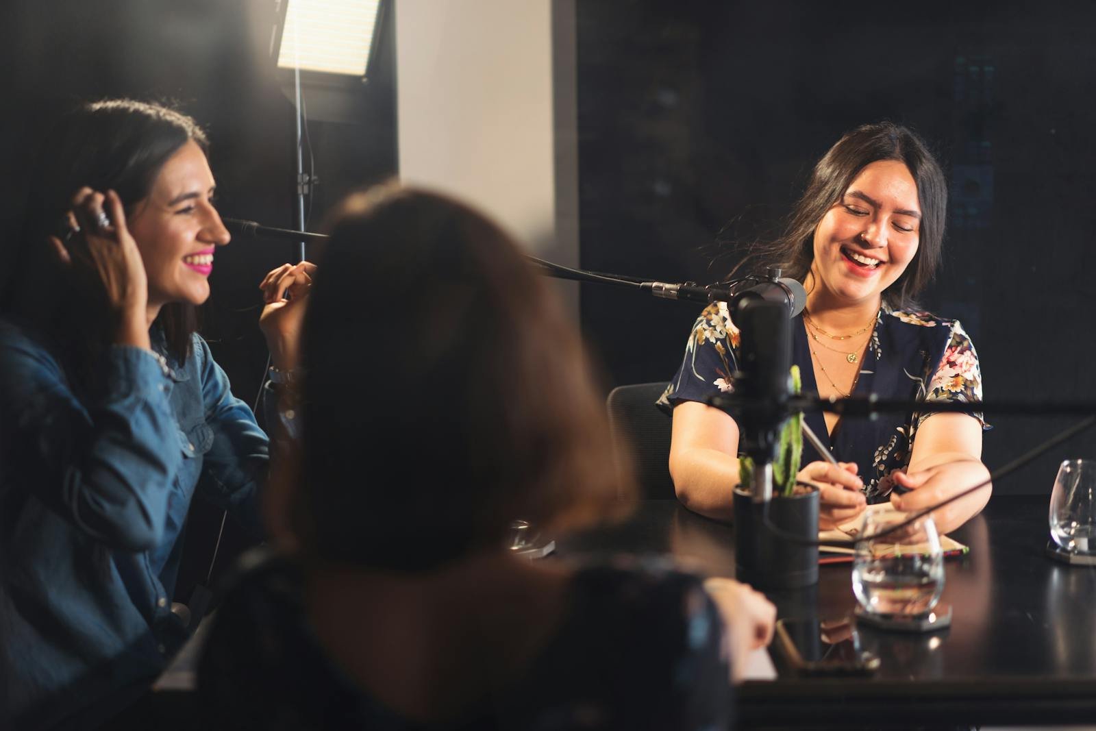 Three women recording a podcast together, sharing smiles and enjoying the moment in a cozy studio setting.