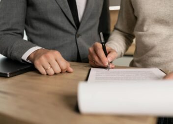 Close-up of businessmen signing documents at a wooden table in an office.