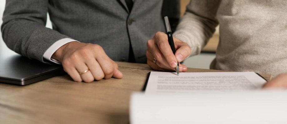 Close-up of businessmen signing documents at a wooden table in an office.