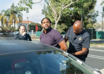 Two police officers detaining a suspect beside a car in a sunny outdoor setting.