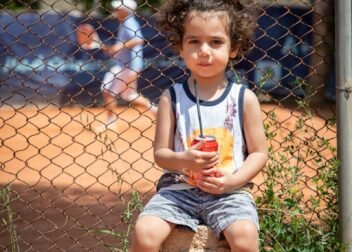 Cute child with curly hair holding a drink sitting outdoors by a tennis court.