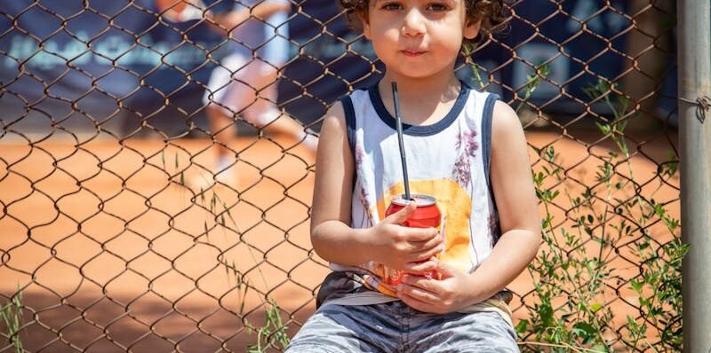 Cute child with curly hair holding a drink sitting outdoors by a tennis court.