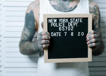 Close-up of tattooed man holding a mugshot sign in front of a height chart.