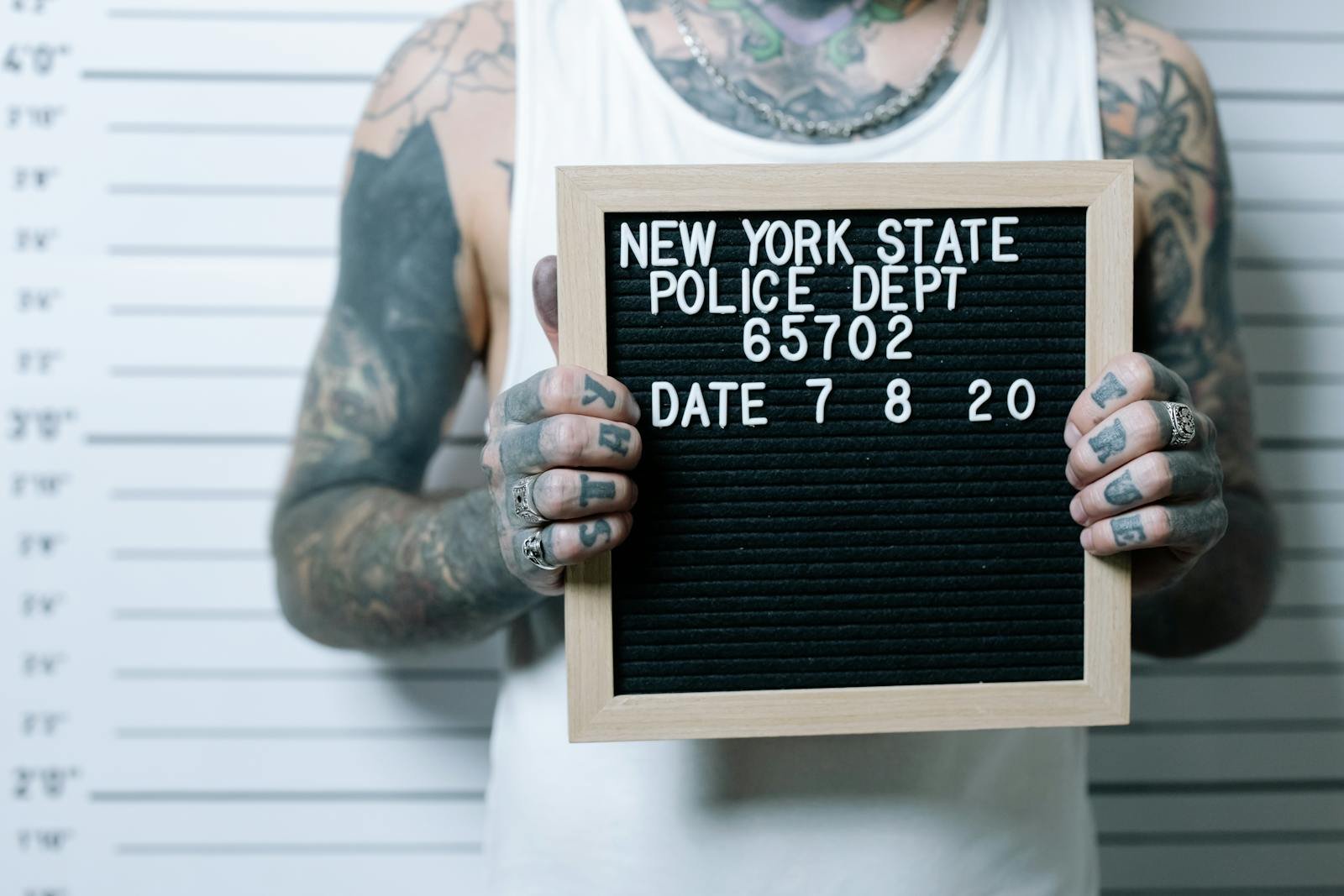 Close-up of tattooed man holding a mugshot sign in front of a height chart.