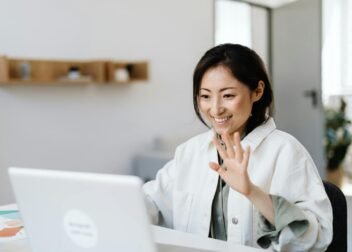 Smiling woman waves at laptop during a video call in a modern office.