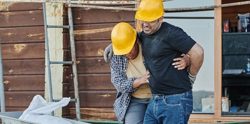 Construction workers wearing helmets assist an injured colleague at a building site with wooden cabins.